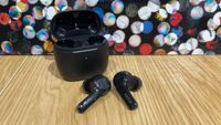 A black pair of Earfun Air wireless earbuds on a wooden surface. The open case, which is also black, is positioned behind them.