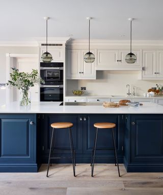 An open plan kitchen with a navy blue kitchen island with white countertops two wooden stools in front of it, white cabinets behind it, and three black cabinets hanging from the ceiling