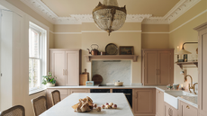 A light pale pink and lemon kitchen with an island and glass chandelier hanging above the white worktop