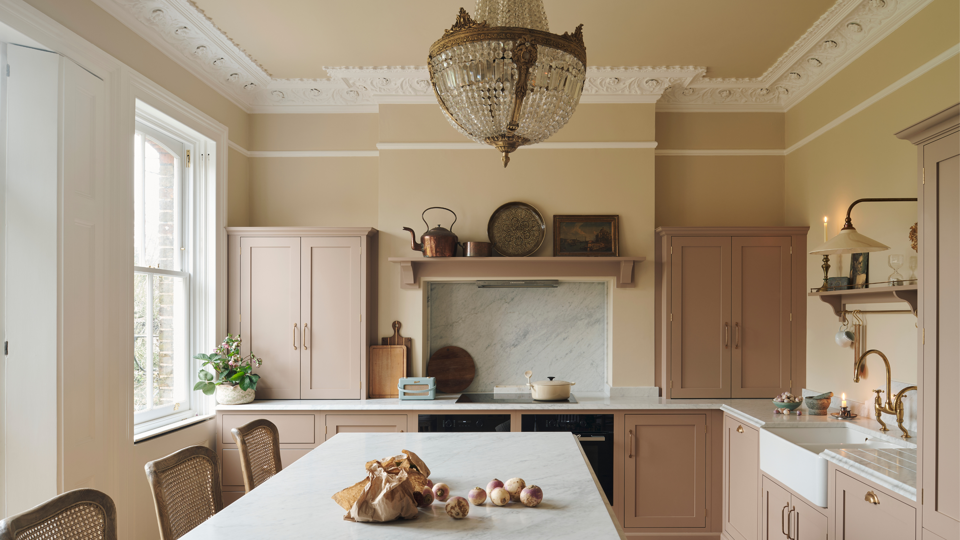 A light pale pink and lemon kitchen with an island and glass chandelier hanging above the white worktop