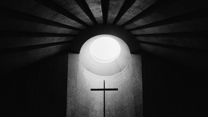 A stark black and white image of a cross illuminated by a circular skylight, casting dramatic shadows and radiating light upwards