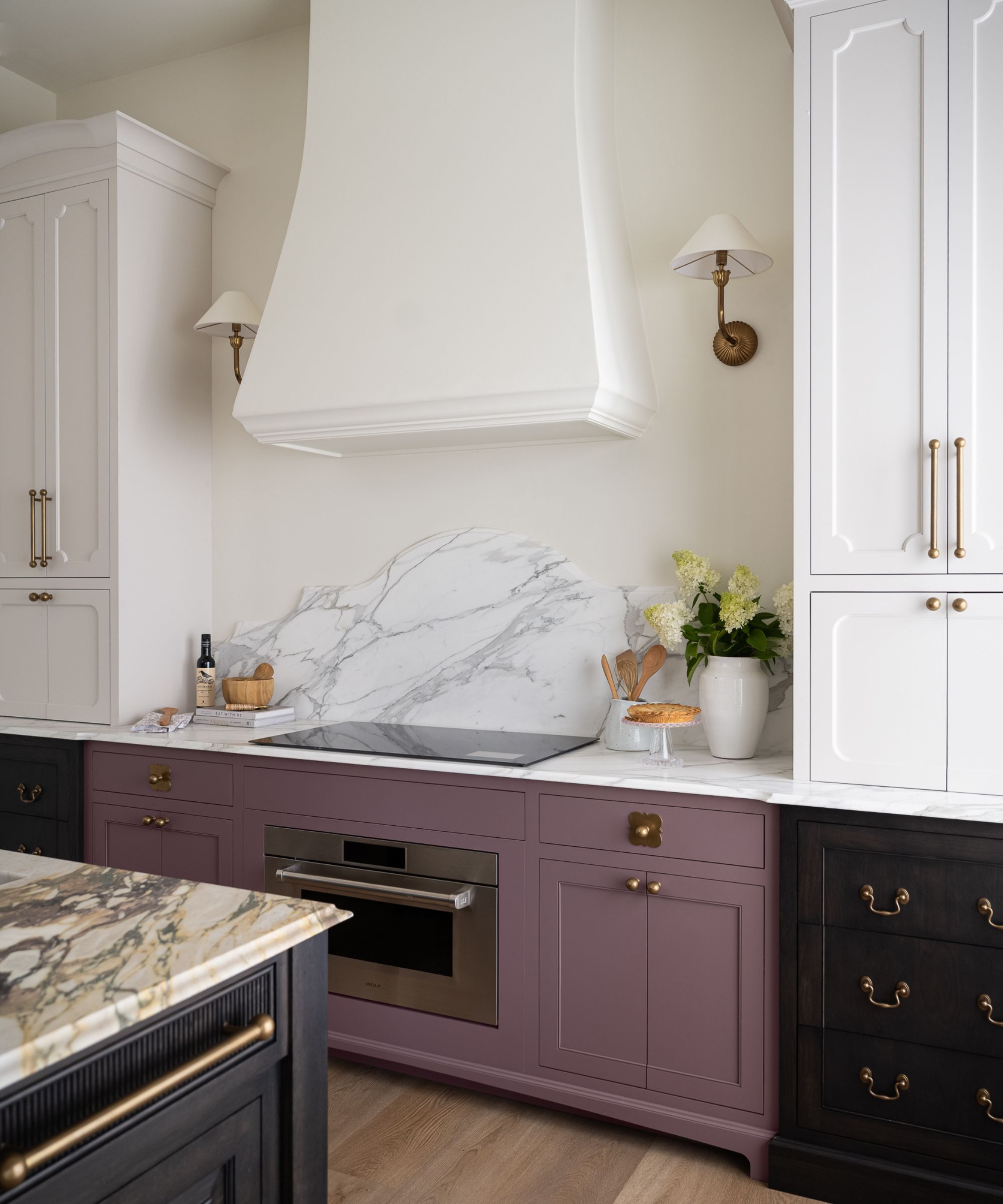 A modern kitchen with white upper cabinets, white walls with two brass wall sconces, a range hood, and a marble countertop. Muted purple lower cabinets with brass hardware and an integrated oven.