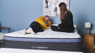 2024's best mattress guide image shows the Helix Midnight Luxe hybrid mattress being tested by our sleep editor and mattress writer, with one lying on the mattress on her side and the other sitting on the edge of the bed to test the edge support