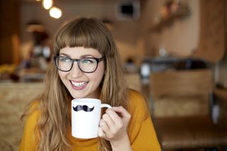 A woman with red hair and glasses is smiling and enjoying a mug of coffee.