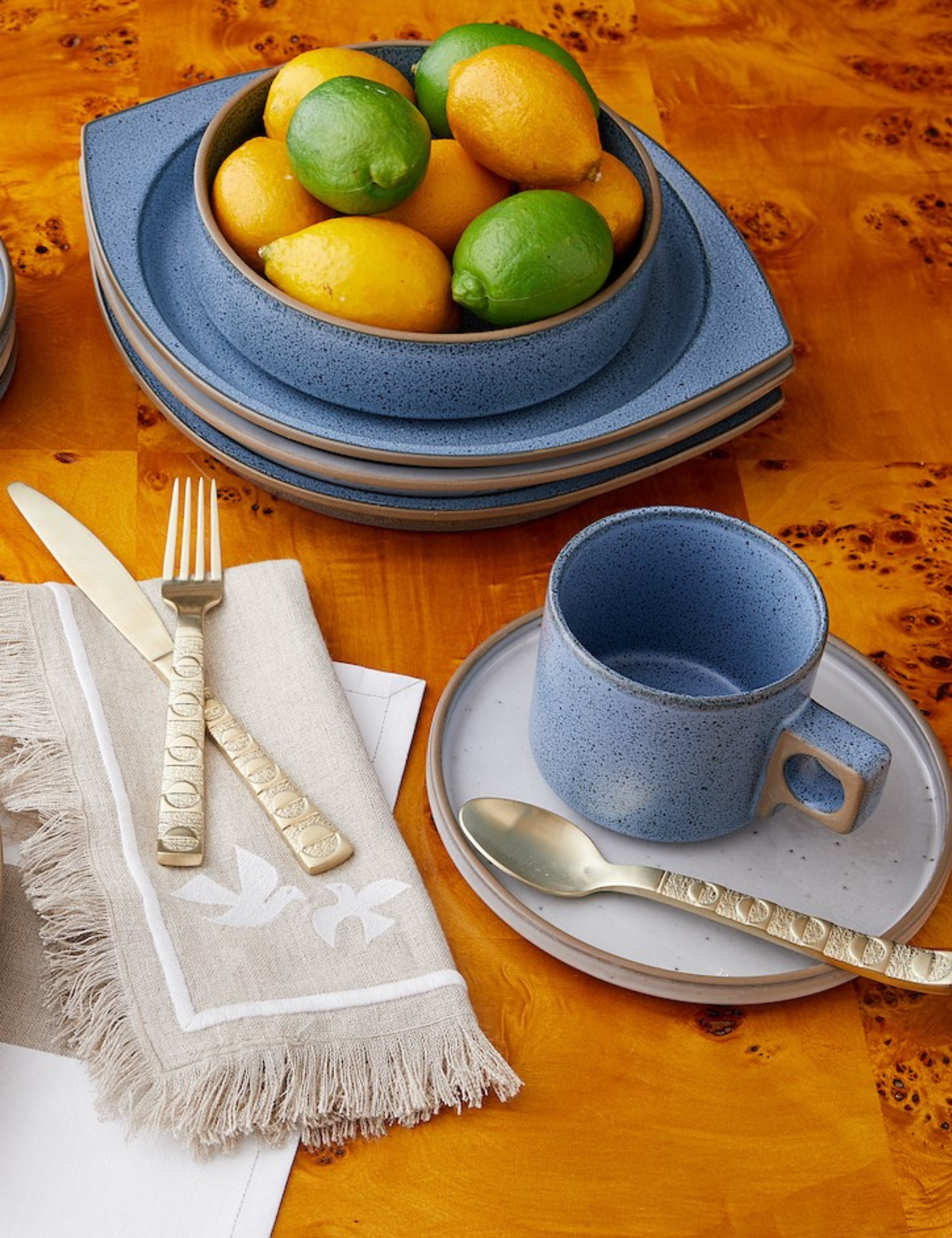 Blue tableware pictured atop of a burlwood dining table alongside sculptural flatware and fringe bordered napkins