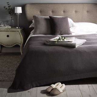 Bed with dark grey and white bedding, slippers and a bedside table