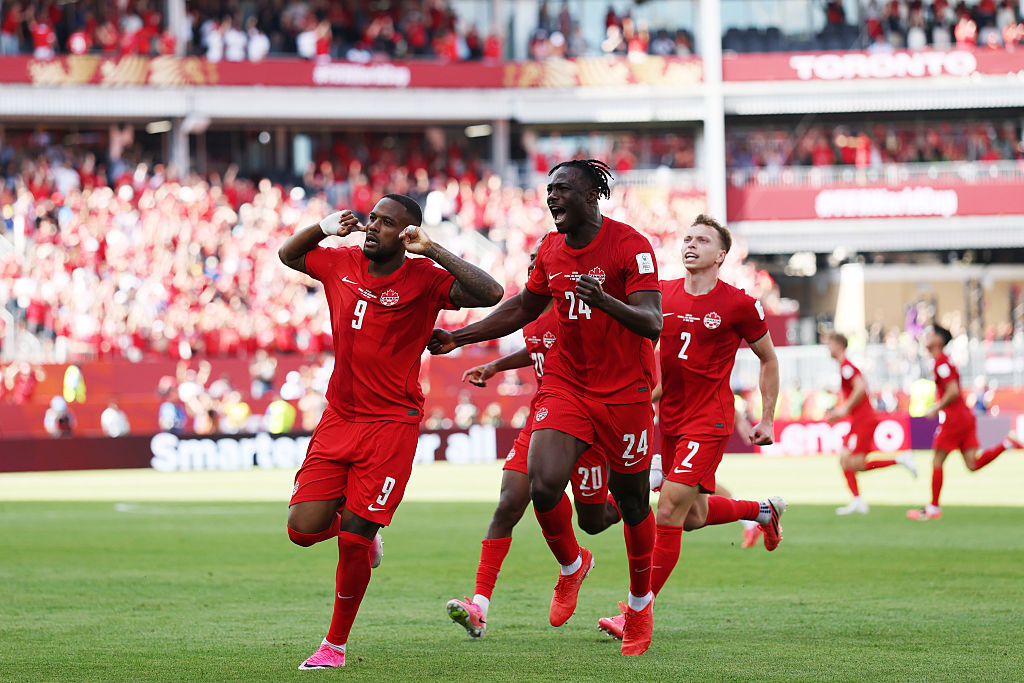 Cyle Larin comemora após marcar pelo Canadá contra a Bósnia, com os dedos pressionados nas orelhas e seus companheiros correndo atrás dele.