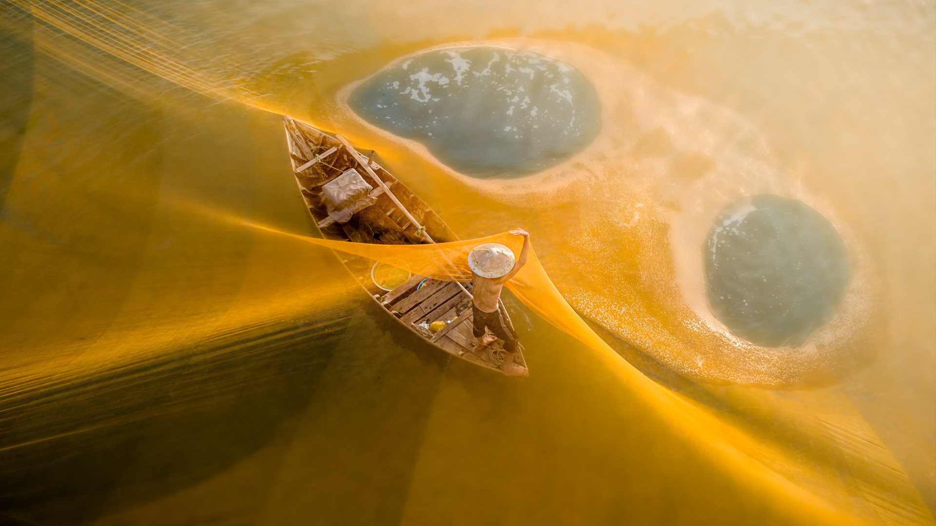An overhead, high-angle shot captures a fisherman in a conical hat standing on a small wooden boat, surrounded by a massive, glowing golden fishing net stretched across the water, which features two distinct, circular clearings.