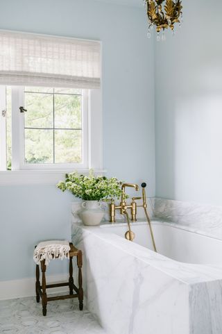 pale blue bathroom with marble tub surround, marble tiled floor, stool, vase of flowers, blind at window, vintage chandelier, gold taps