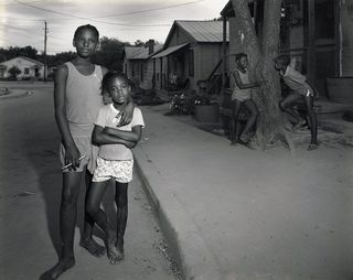 A black-and-white photo depicts four children, two standing on the left in sleeveless tops and shorts, and two talking while leaning against a tree in a industrial neighborhood.