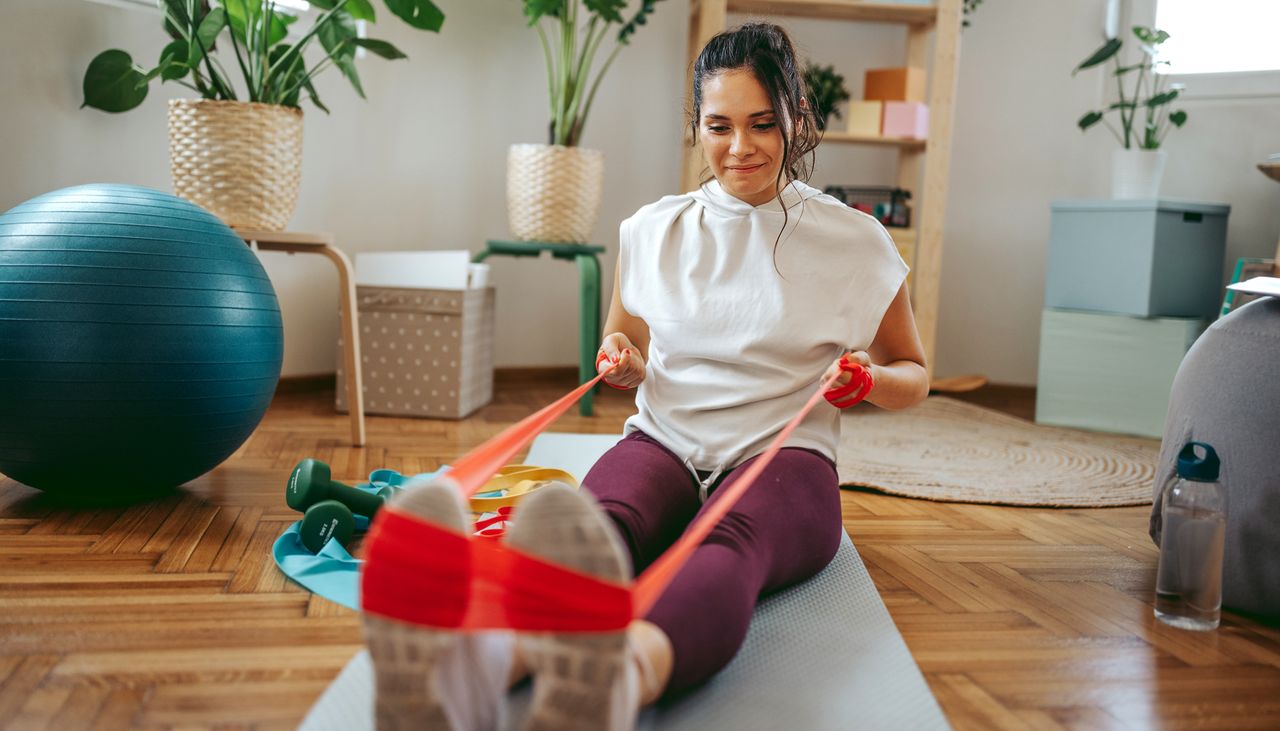 Woman exercises at home using a resistance band. She is sitting on the floor with her legs extended in front of her, holding a resistance band wrapped around her feet,