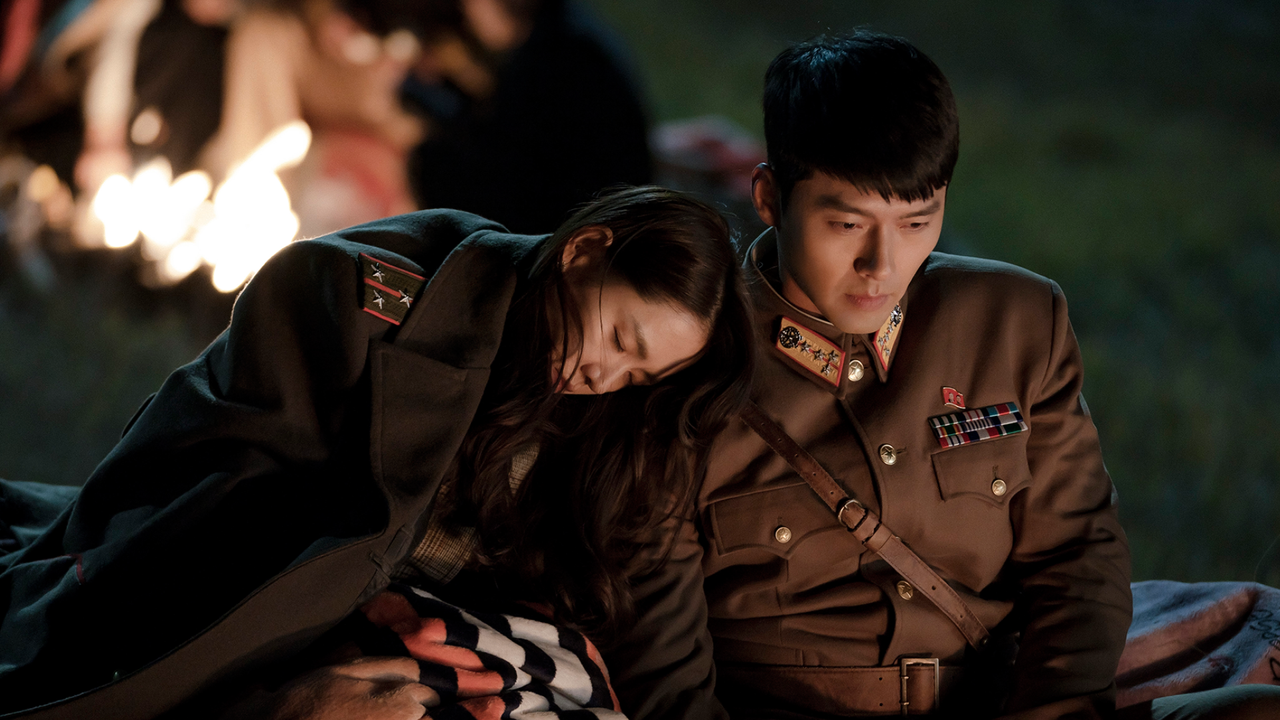 a woman wears a military jacket and rests her head on a soldier&#039;s shoulder outside of a fire in a still from crash landing on you