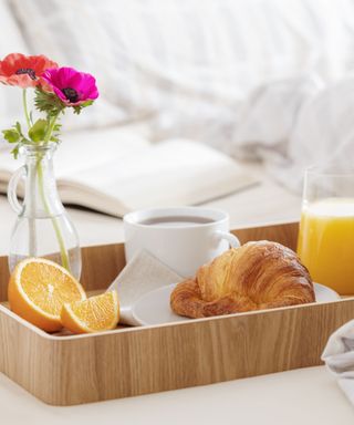 A wooden tray with oranges, coffee cups, a corissant, orange juice and a vase of pink flowers on it on a white bed