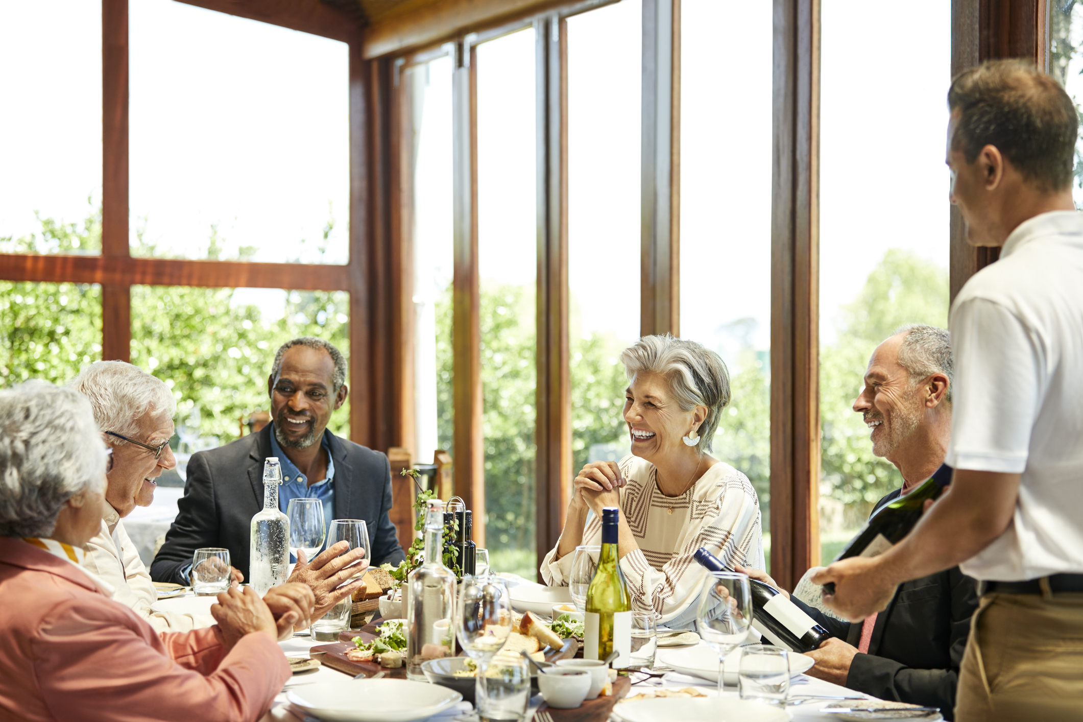 Male sommelier with wine bottle standing by smiling senior friends. Men and women are sitting in restaurant. They are spending leisure time.