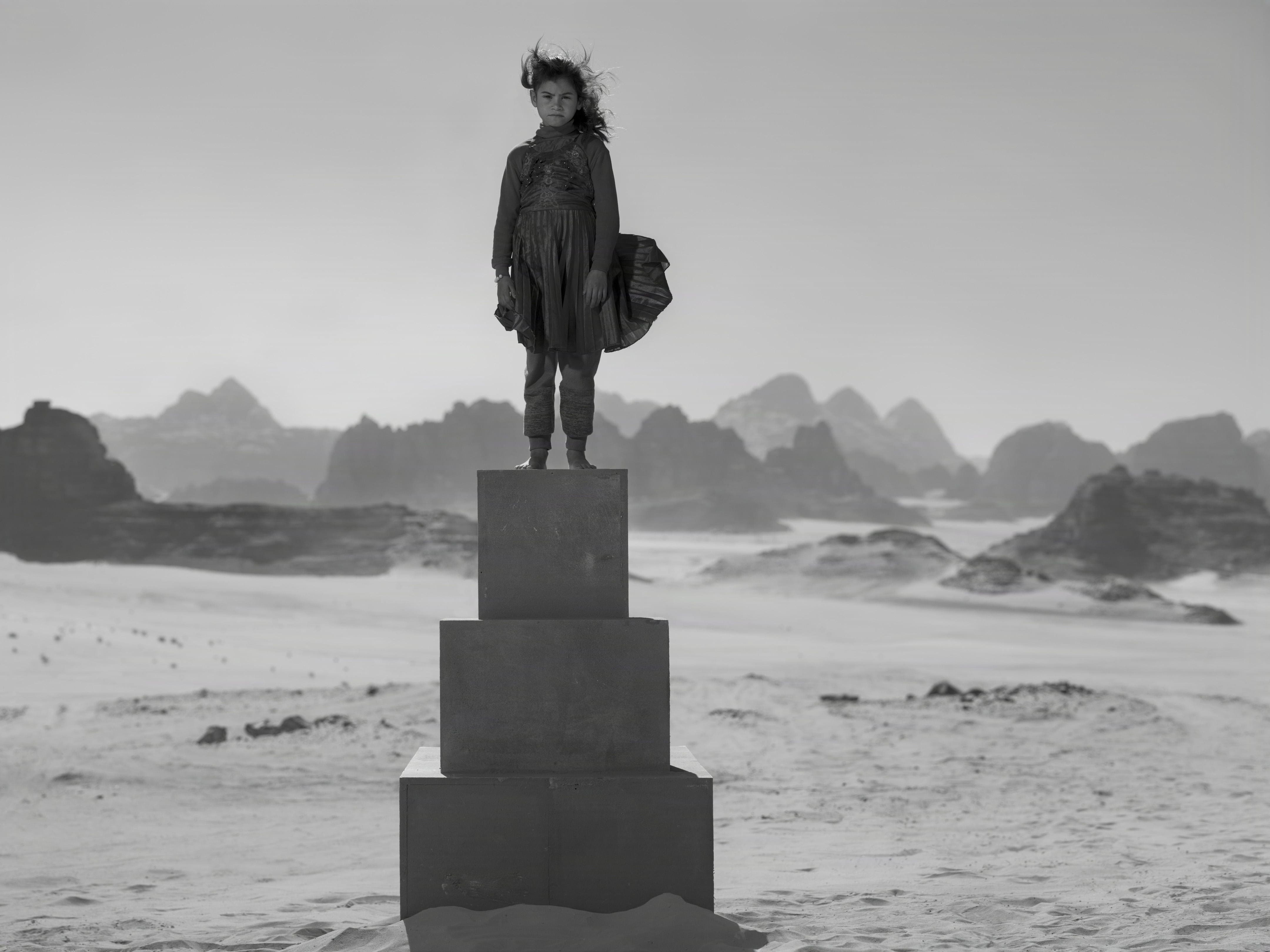 A young girl stands defiantly atop a tall pedestal of stacked blocks in the middle of a barren desert.
