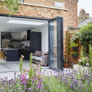 Exterior view of kitchen extension, brick wall and folding glass doors with black metal frames, backing onto a garden filled with bee friendly flowers