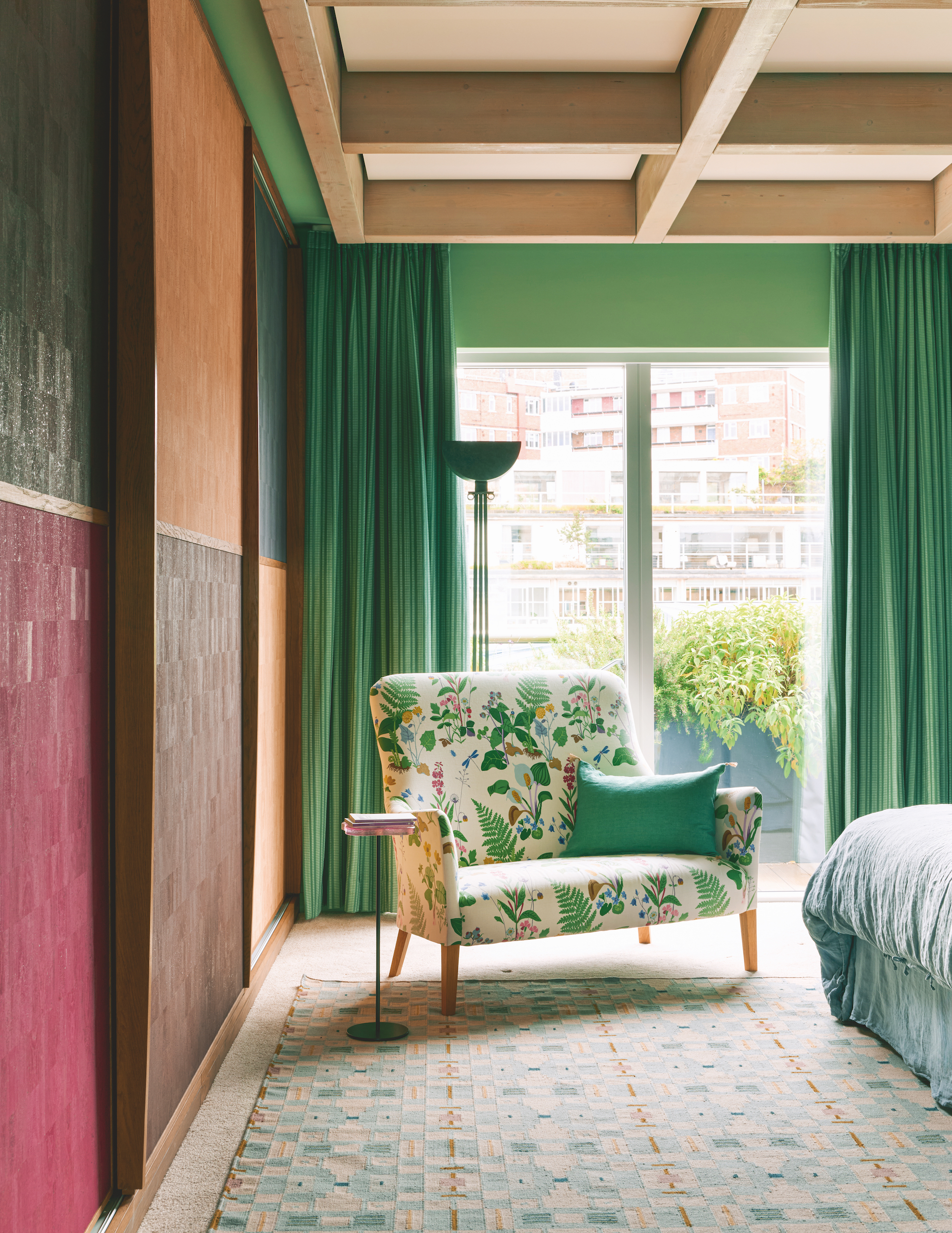 A modern bedroom with striking green curtains on the large window at the back of the space, there is a printed armchair just in front with a lamp behind it, the sliding door to the wardrobe on the left are multi colored and there is the bed in front of that.