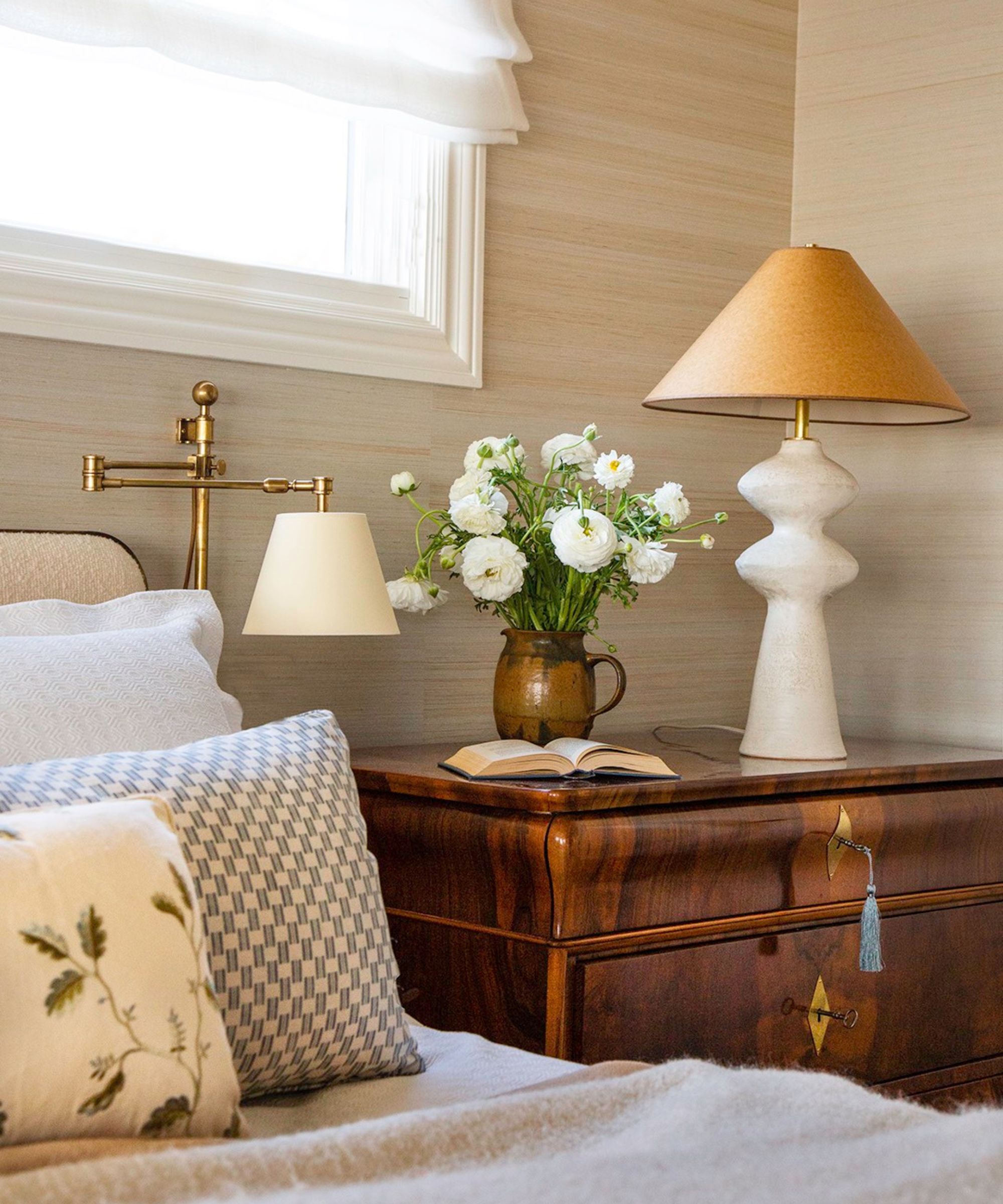 Bedroom with beige grasscloth walls, antique dresser, lamp, vase of flowers, gold sconce and bed with blue and floral cushions