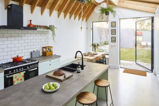 Cut extension costs: a kitchen with large pivot door and picture window, concrete-effect worktops and floor, and exposed beams in the pitched extension roof