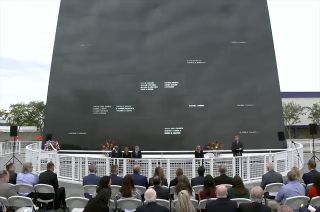 a handful of people in dark suits stand in front of a large black wall into which several dozen names have been carved.