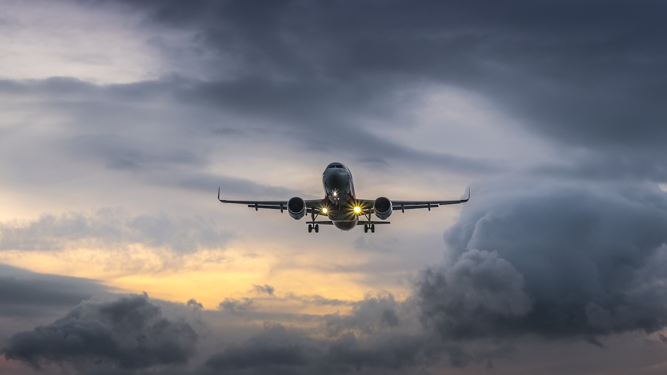 plane landing during sunset, there are dark stormy looking clouds behind it.