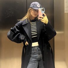 French editor and writer Eugenie Trochu poes for an elevator mirror selfie wearing a trench coat with a baseball cap