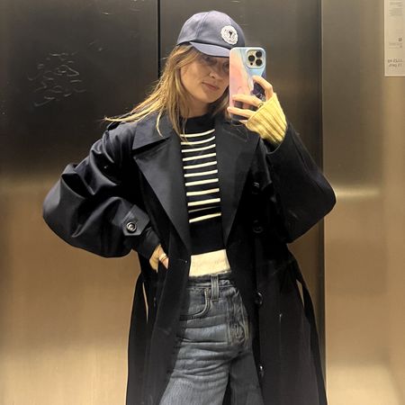 French editor and writer Eugenie Trochu poes for an elevator mirror selfie wearing a trench coat with a baseball cap