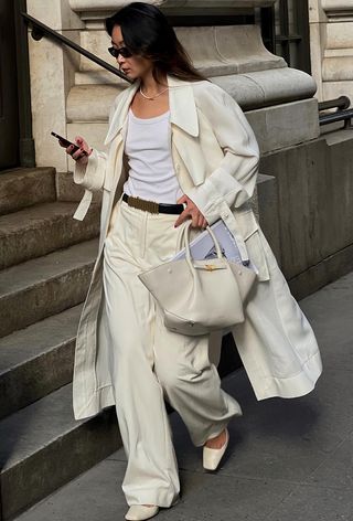 a photo showing what to wear to work in the summer with a woman wearing a white trench coat over a white tank top with white trousers, white pumps, and a white structured tote bag