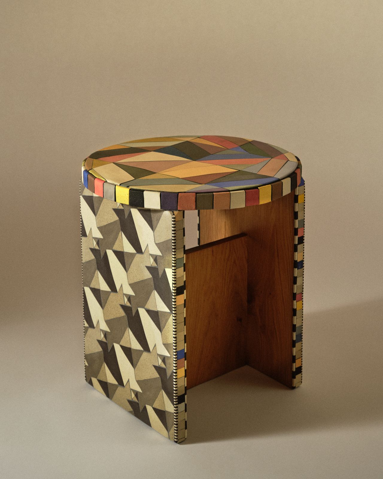 A wooden stool boasts a surface entirely covered by geometrical triangular shapes in tones of blue, yellow, red, grey, white, black, brown, and beige.