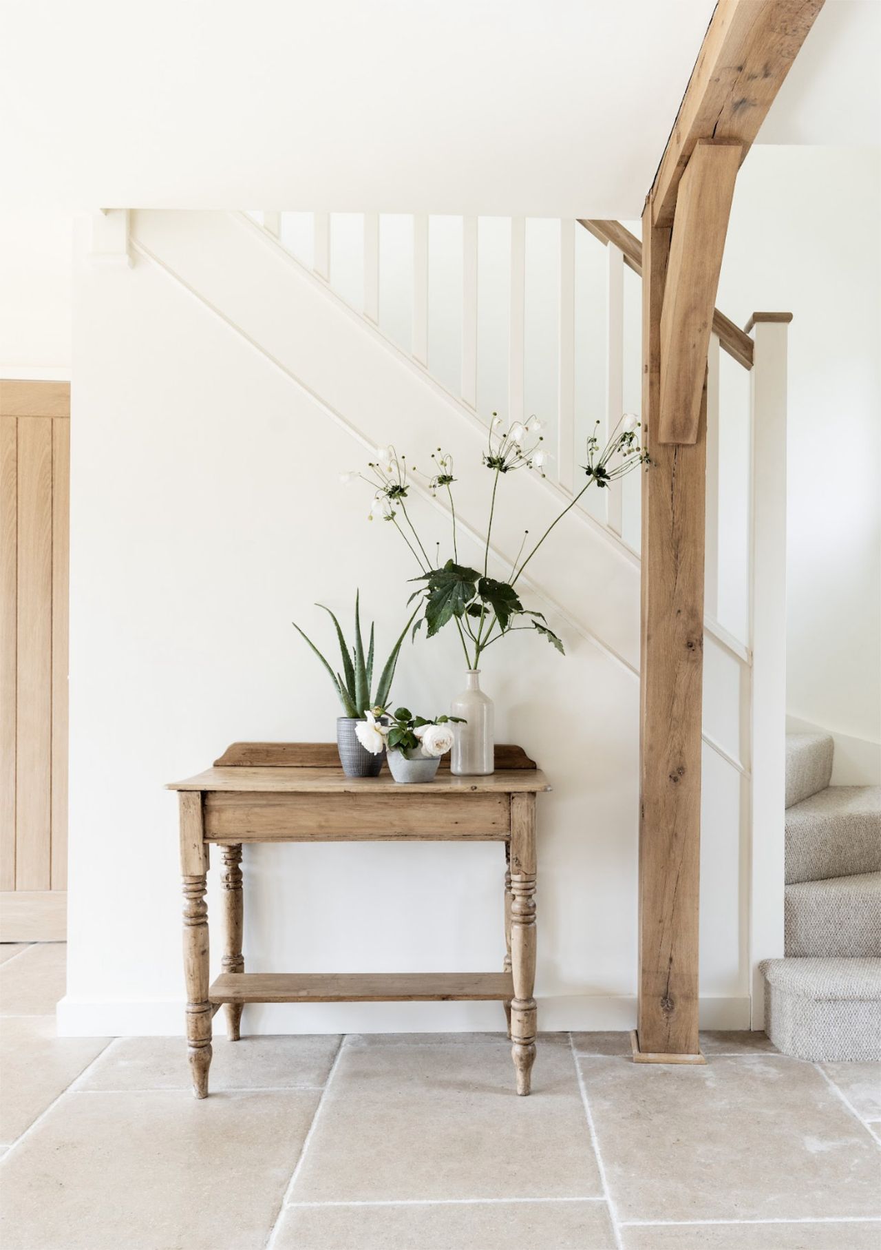A white hallway with light flagstone floor tiles, a wooden console table topped with flowers, and a stairway leading to a second floor