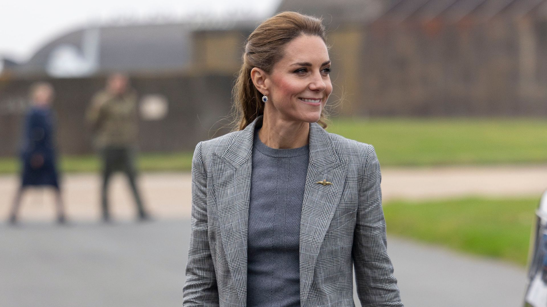 Kate Middleton walks outside wearing a tonal grey suit with navy suede heels and a black leather belt.