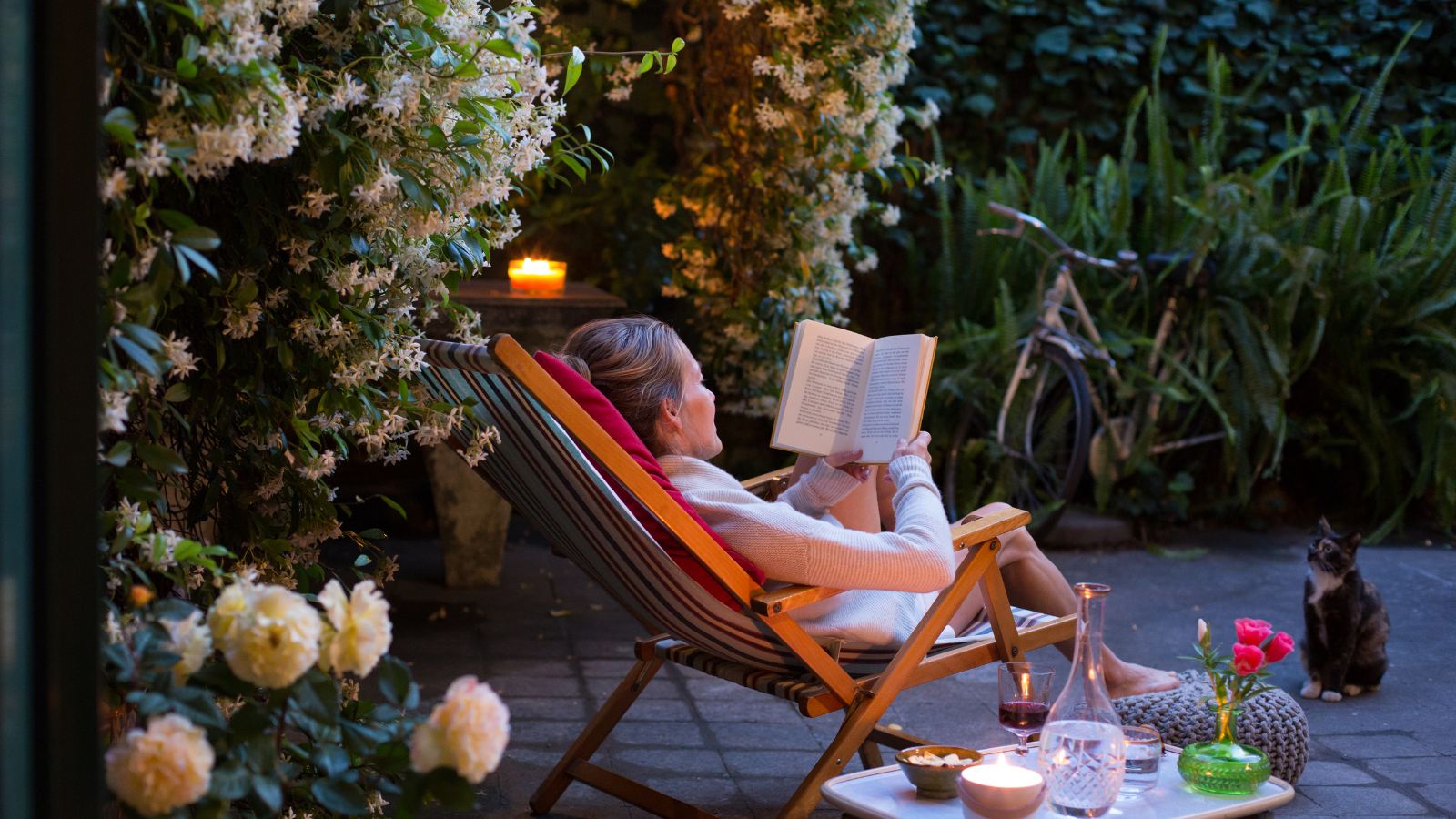 Woman relaxing on deck chair in back yard, reading a book with her cat watching 
