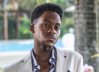 Hugo Kingsley (Tyrone Huntley) pictured at the hotel's outdoor restaurant with the pool visible in the background. He has a self-satisfied look on his face.