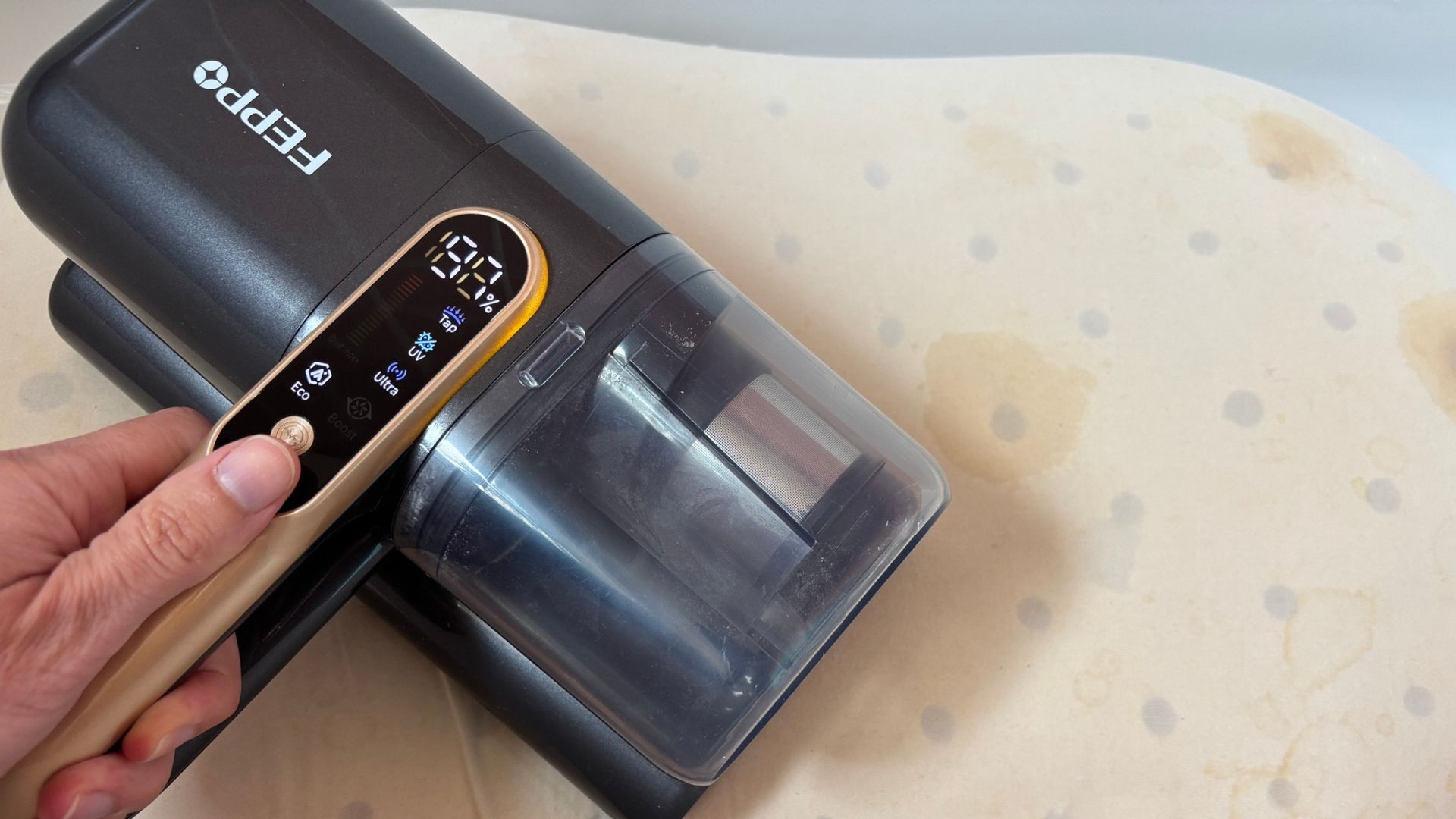 The image shows a hand gripping a cordless mattress vacuum, which is moving over the surface of a stained memory foam pillow.
