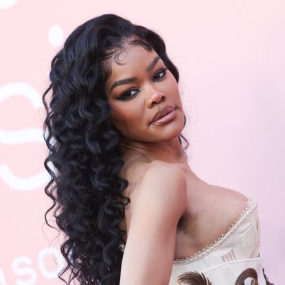 a black woman with long, curly hair poses in front of a pink backdrop at a red carpet event