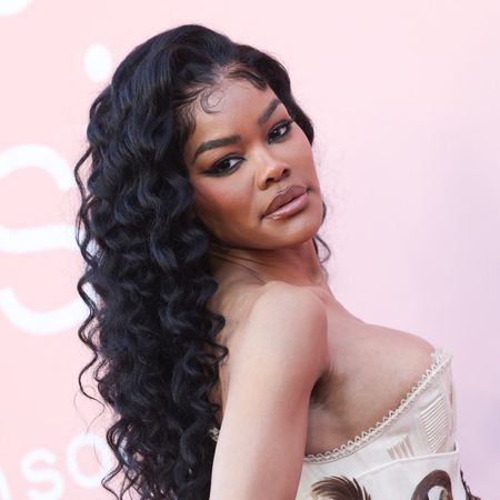 a black woman with long, curly hair poses in front of a pink backdrop at a red carpet event