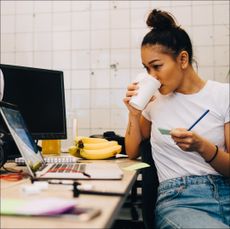 A woman drinking a coffee and working at a desk with a computer