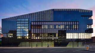 A photo of the Graphene Institute at the University of Manchester, taken from side-on. Decorative: the building was photographed at night, with evening sky seen behind and lights on inside the building.