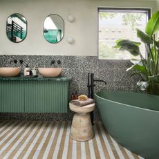 bathroom with beige and white striped floor tiles, a large green bathtub, an oversized banana plant and a green fluted bathroom console unit with twin pink/beige basins and black hardware