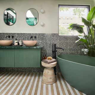 bathroom with beige and white striped floor tiles, a large green bathtub, an oversized banana plant and a green fluted bathroom console unit with twin pink/beige basins and black hardware