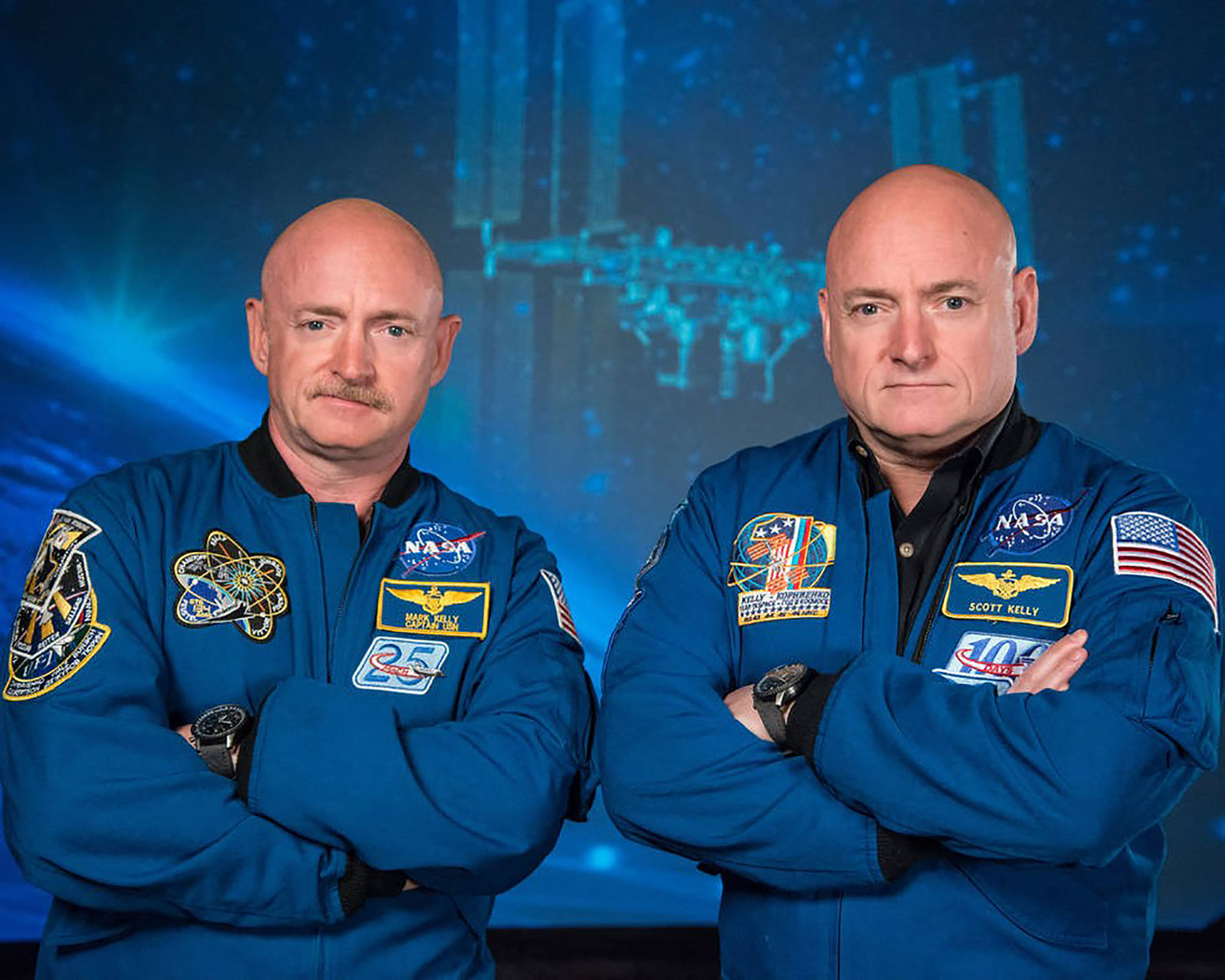 a pair of twins, Scott and Mark Kelly sit with their arms folded wearing NASA flight suits.