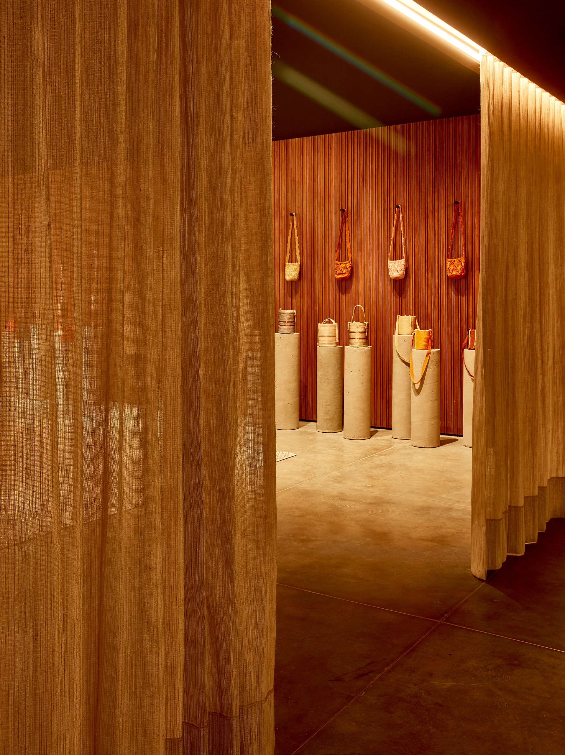 An orange-tinted showroom of textiles in natural fibers features a series of cement pillars on top of which sit handbags in sculptural sizes, with more bags hung up on the curtain background.