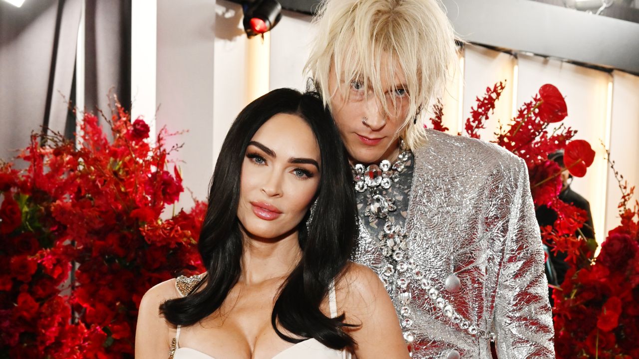 Megan Fox is smiling and has long black hair while Machine Gun Kelly stands behind her with shaggy blonde hair and wearing a silver suit