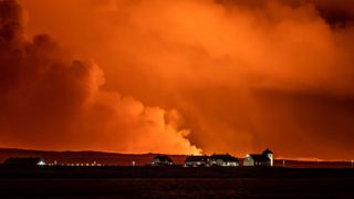A view of the Bessastadir, four buildings in the distance with lights on, the official residence of President of Iceland as volcano spews smoke behind.
