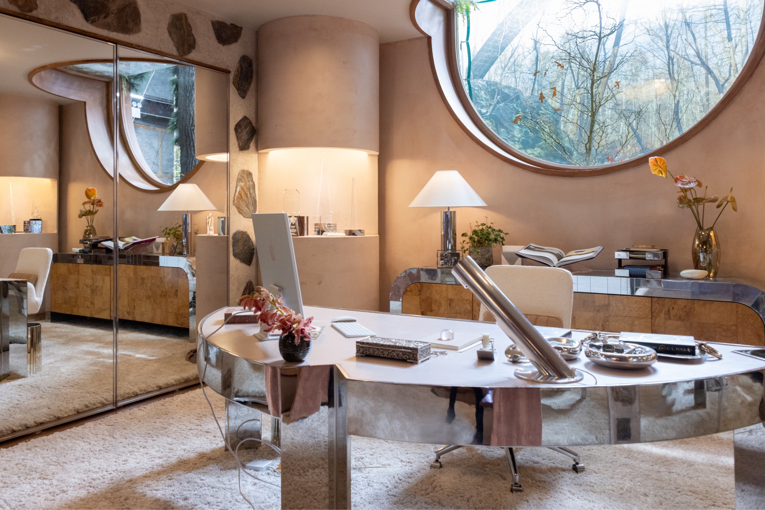 A pale pink, mid-century office with a cream rug covering its floor, a semicircular desk with a mirrored set of legs and body and a cement surface, warm soft lighting, and a semi-circular window.