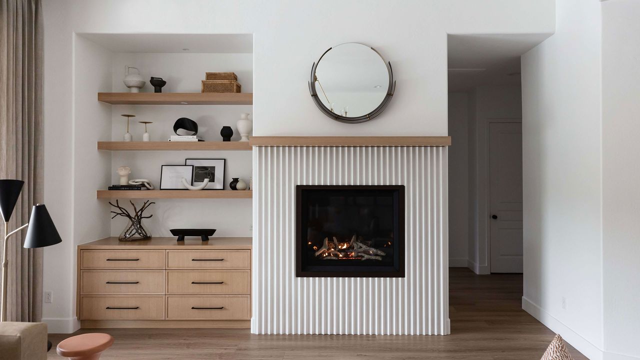 Round ornate mirror above modern fireplace in white living room with open shelving