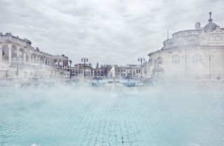 Thermal waters of Budapest. Large outdoor baths with blue water surrounded by mist and historical buildings.