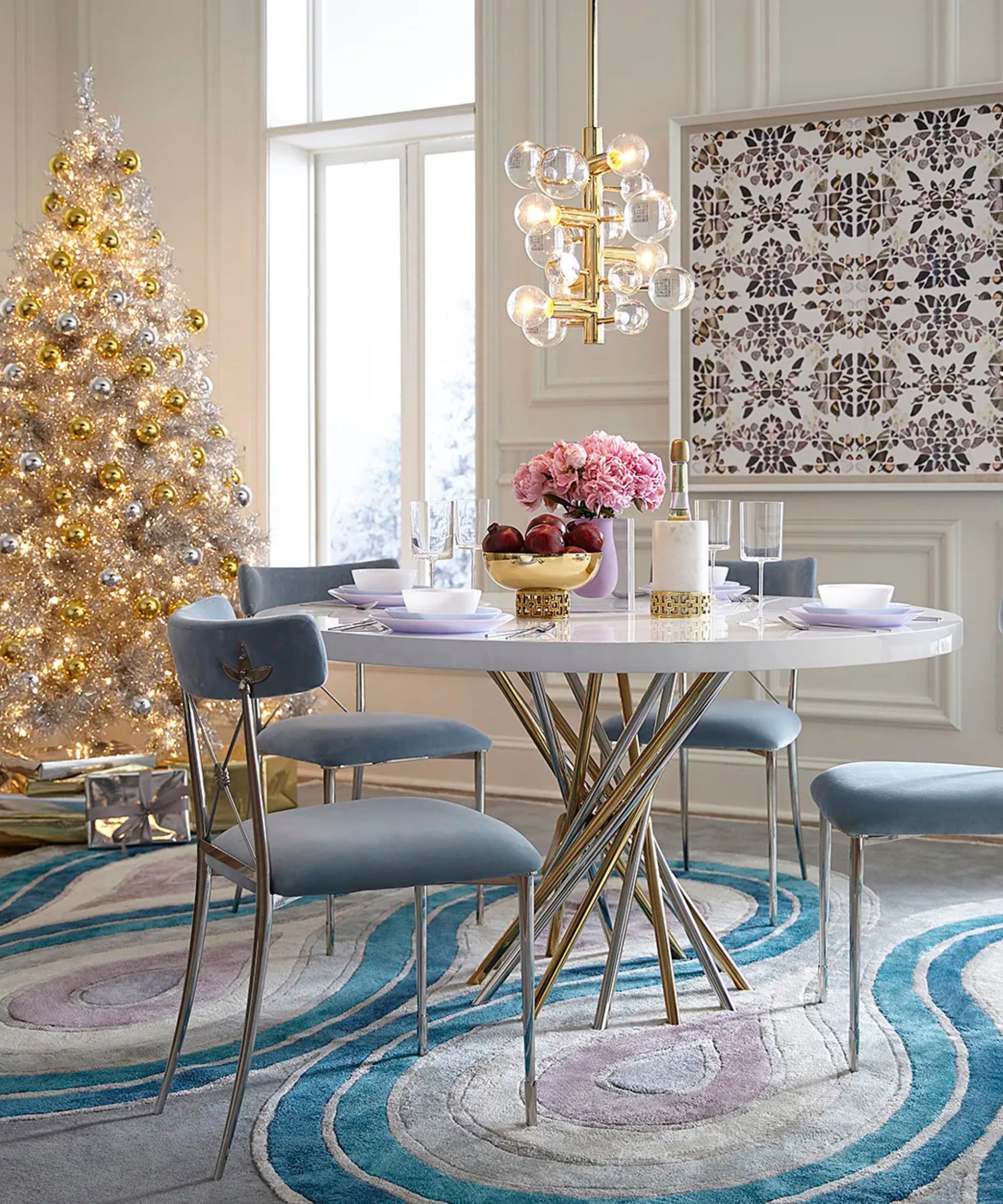 A bright dining room with a white marble table, blue velvet chairs, and a shimmering silver tinsel Christmas tree decorated with gold ornaments.