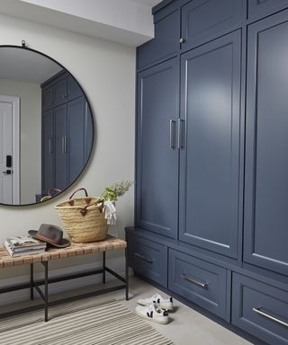 A hallway shoe storage idea with blue cabinets, shoe bench and large circular mirror