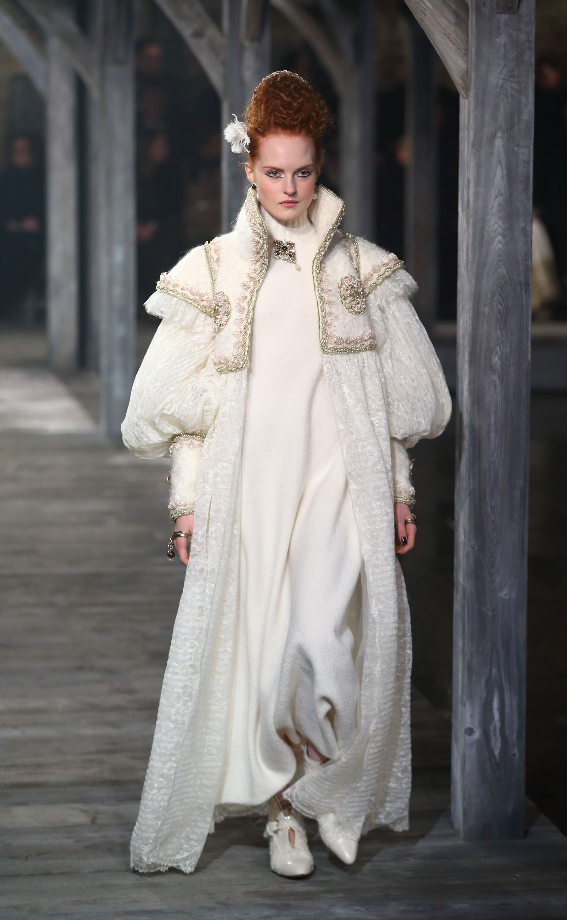 Red-haired model in ivory coloured robes walking down a wooden runway